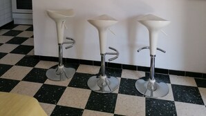 Een keuken met een geruit vloertje, een houten aanrecht, een spoelbak, een fornuis en drie witte barstoelen.