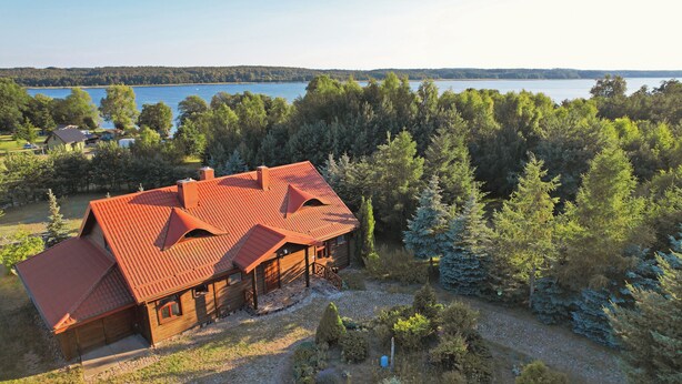 A wooden cabin with a red roof, surrounded by dense forest and a lake in the background.