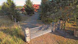 A rustic wooden gate leading to a stone pathway, surrounded by dense evergreen trees and a red-roofed building in the background.