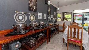 Una mesa de madera con una variedad de platos y utensilios para un bufé, un refrigerador lleno de bebidas, y una ventana que da a zonas verdes.