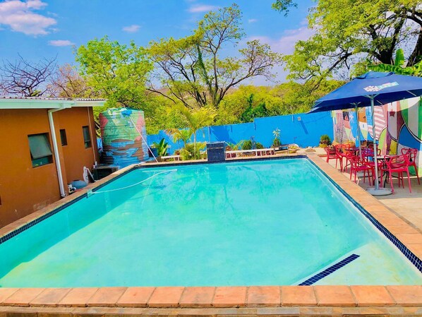 A swimming pool with a tiled edge, surrounded by a brick border. There are red chairs and a blue umbrella under a mural. - Woodpeckers Guest House (Mbombela)
