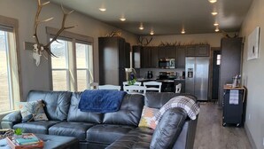 Ein modernes Wohnzimmer mit einer Ledersofa, einem Holzschemel und einem Couchtisch. Im Hintergrund ist eine Küche mit dunklen Schränken und Edelstahlgeräten zu sehen.