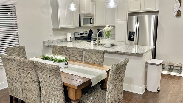 A modern kitchen with white cabinetry, stainless steel appliances, and a dining area with wicker chairs and a wooden table. - Relax Near Beach, Shops, Parks, Mayo Clinic, Quick Drive to Downtown & Stadium (Jacksonville Beach)