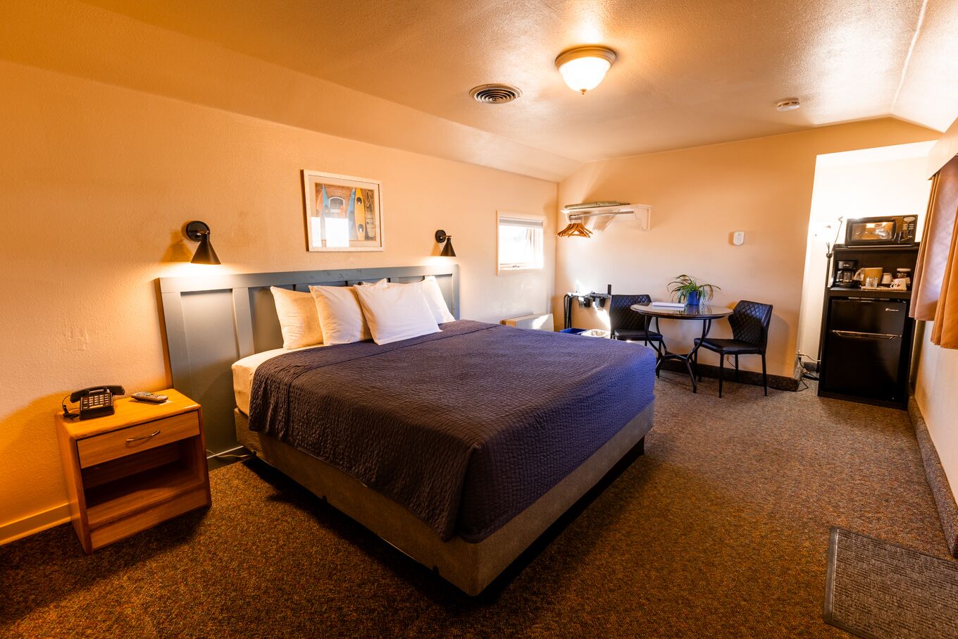 A hotel room with a large bed, bedside table with a phone, a desk with a chair, and a kitchenette.