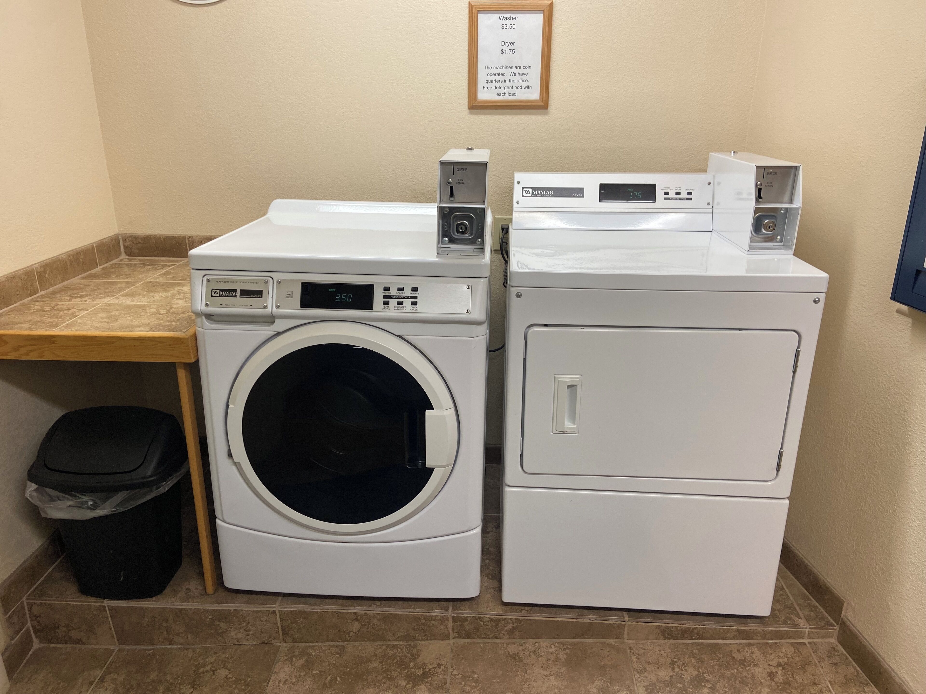 A laundry area with two washing machines and a dryer, a trash bin, and a wooden counter.