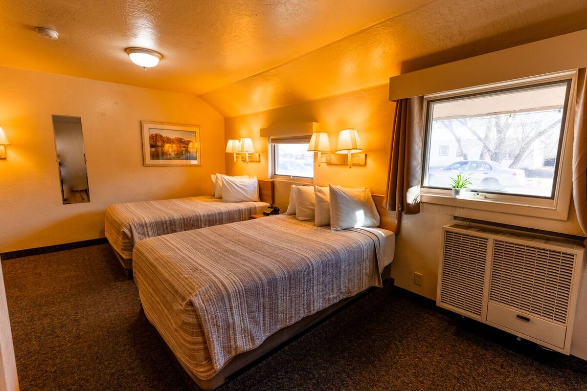 A hotel room with two beds, a window with a view, a heater, and a painting on the wall.