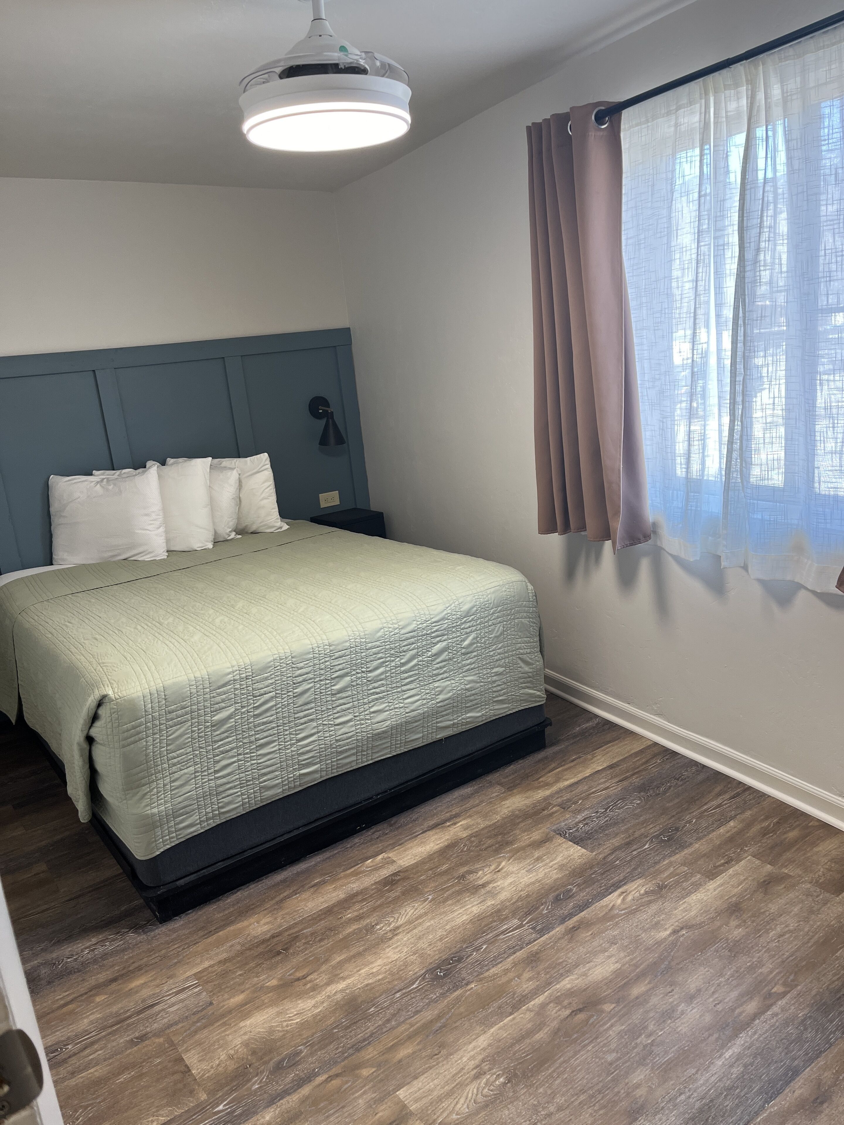 Un dormitorio con cama, mesita de noche, lámpara, luz de techo y ventana con cortinas.