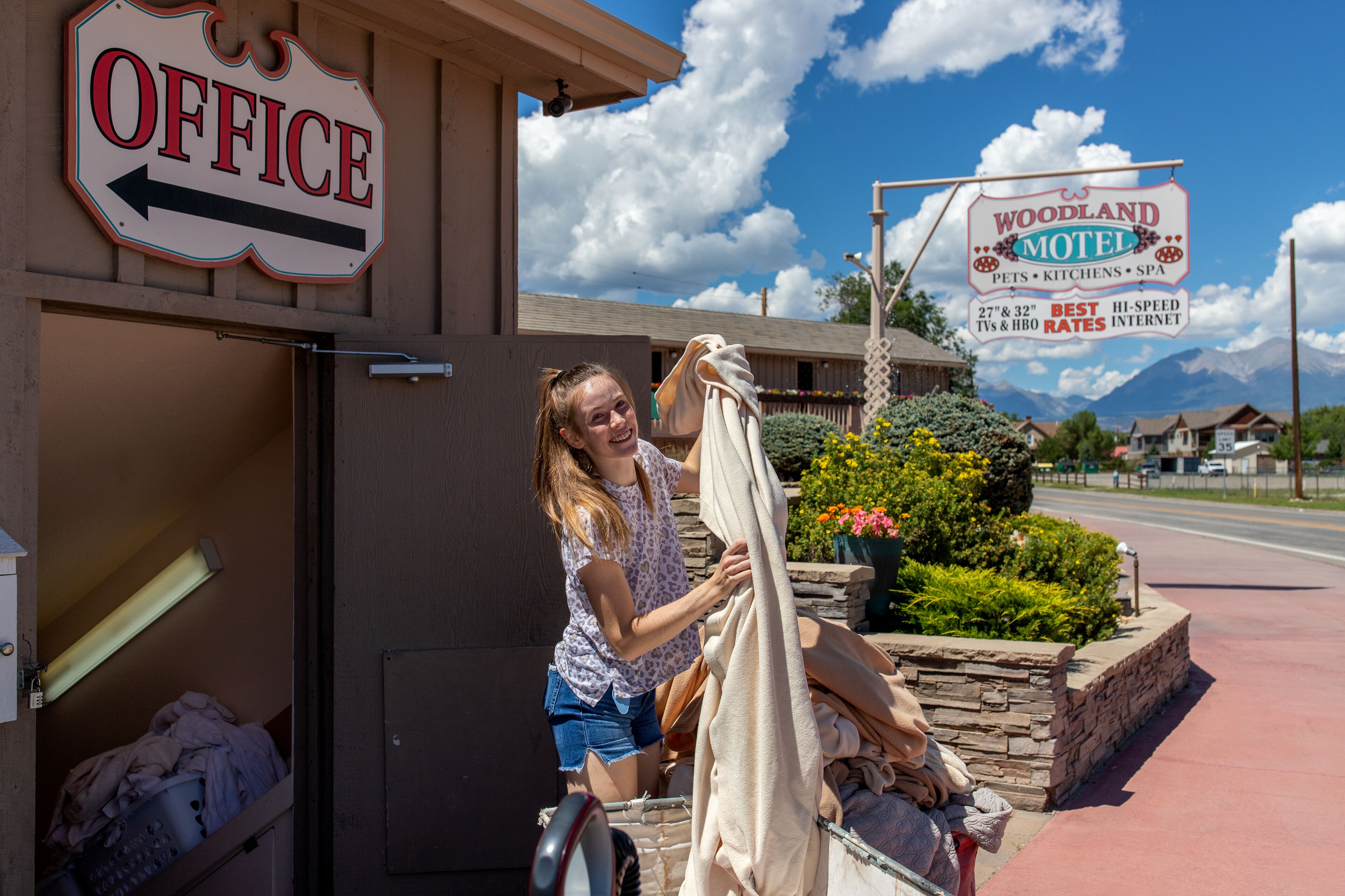 A motel sign with amenities listed, a laundry cart outside, and a clear sky.