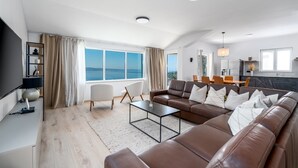 A modern living room with a large brown leather sofa, a coffee table, and a TV on a black stand. There is a large window with curtains, a dining area with chairs, and a kitchen in the background. - New! Villa Atopos with heated private pool, 5 bedrooms, Cinema room, panoramic sea views (Omis)