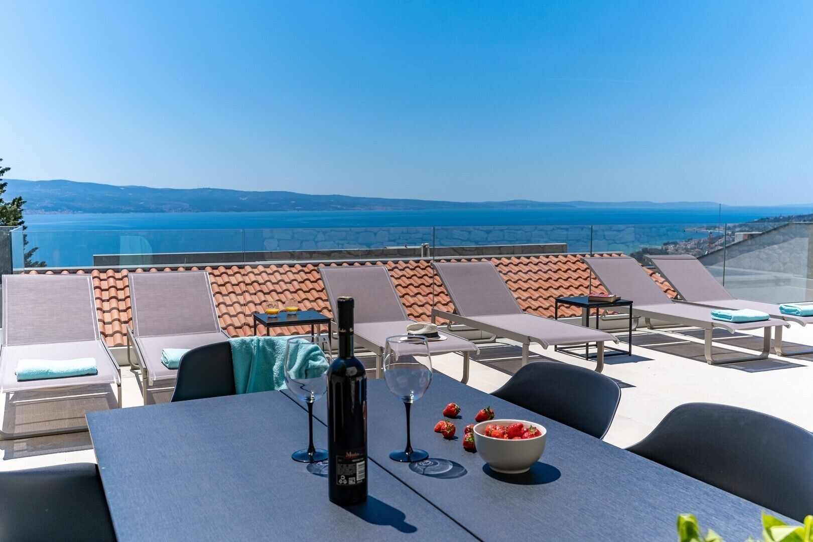 A rooftop terrace with lounge chairs, a table with a bottle of wine, glasses, and a bowl of strawberries.