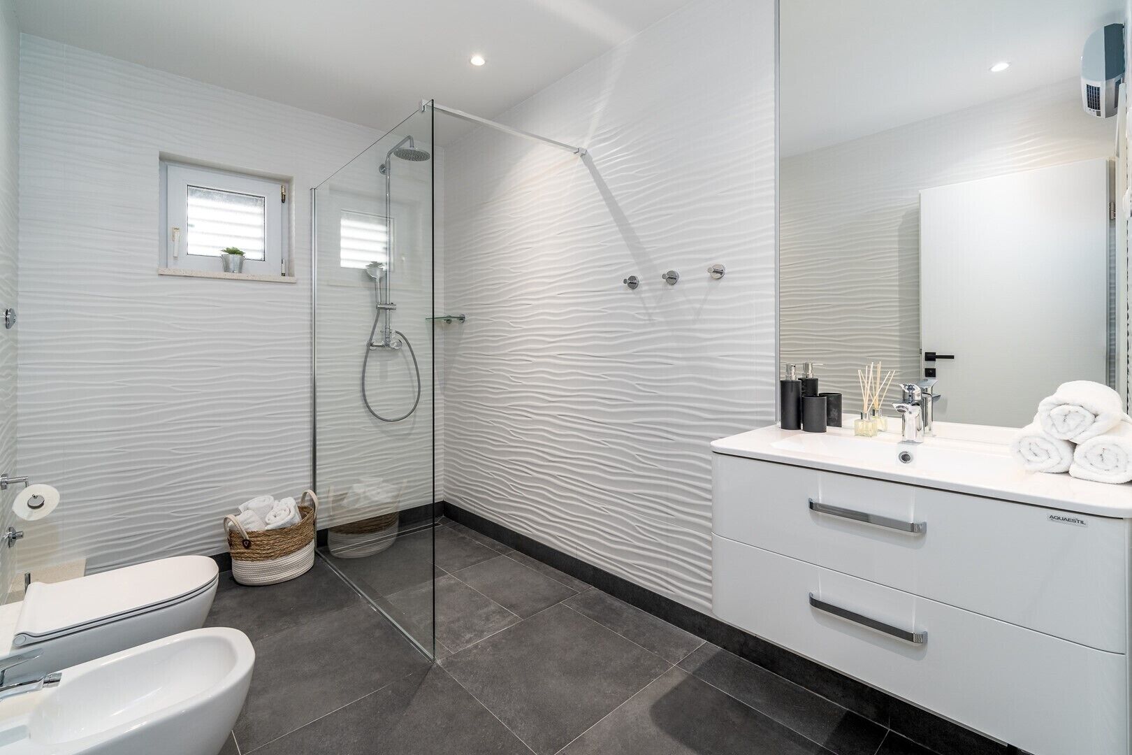 Une salle de bain moderne avec une douche à l’italienne en verre, un meuble-lavabo blanc doté d’un grand miroir et un sol carrelé foncé.
