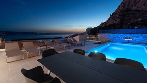 A rooftop pool area with lounge chairs and a table, overlooking a coastal landscape at dusk. - New! Villa Atopos with heated private pool, 5 bedrooms, Cinema room, panoramic sea views (Omis)