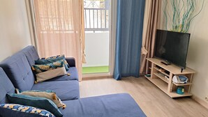 Een woonkamer met een blauwe bank, een televisie op een houten dressoir en uitzicht buiten door een glazen deur.