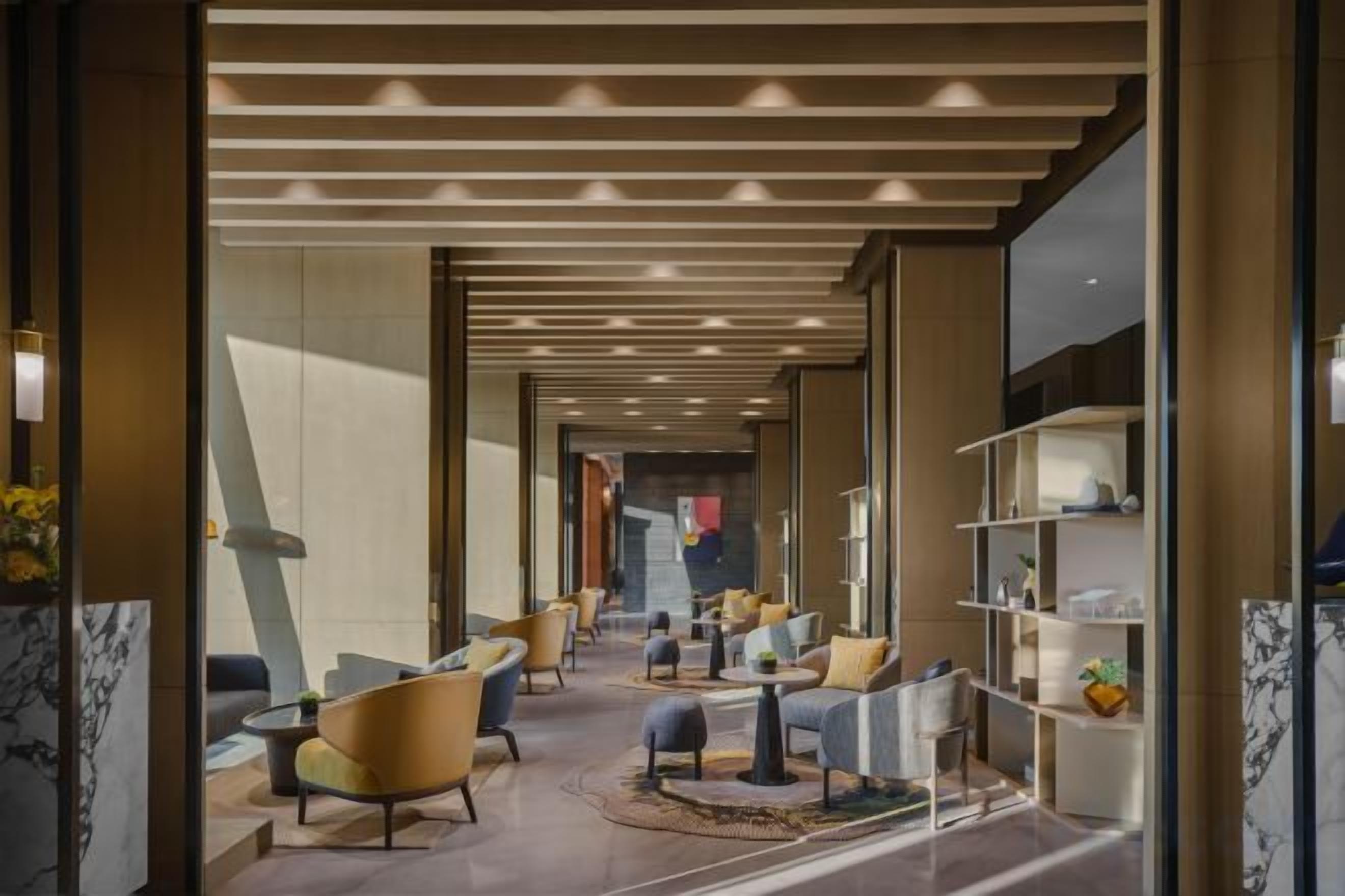 Een hotel lobby met een modern design, met een centraal zitgedeelte met banken en fauteuils, een marmeren receptiebalie en een glazen scheidingswand met planken.