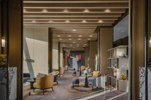 Um saguão de hotel com design moderno, apresentando uma área de estar central com sofás e poltronas, um balcão de recepção de mármore e uma divisória de vidro com prateleiras.