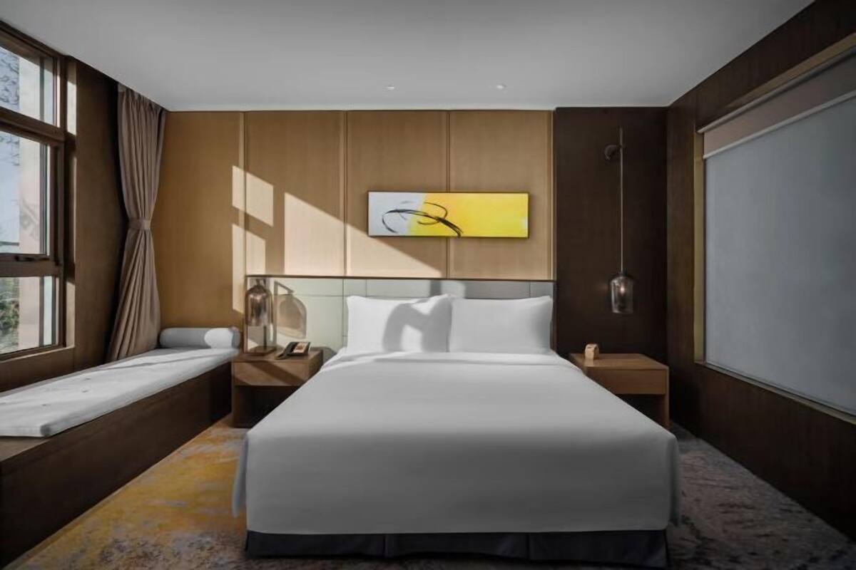 Een hotelkamer met een bed, nachtkastje, gordijnen voor het raam en een kunstwerk aan de muur.