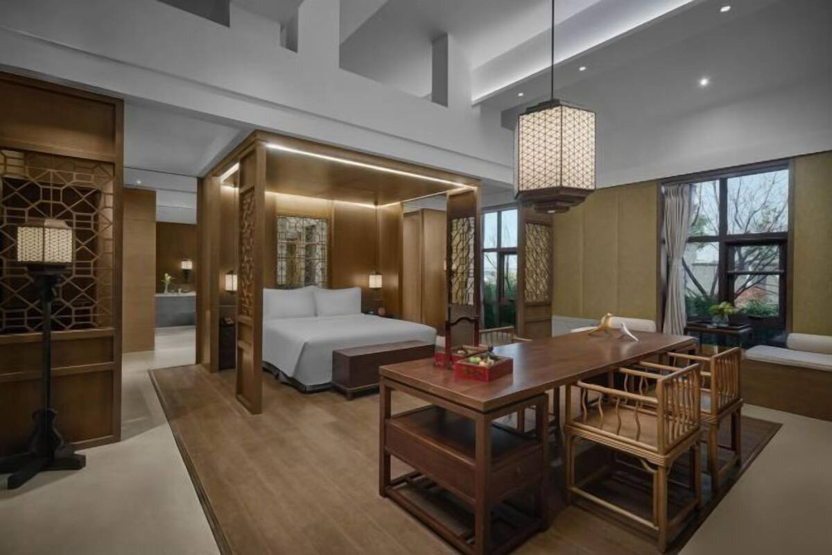 Een moderne hotelkamer met een houten vloer, een bed, een eettafel met stoelen, een hanglamp en een groot raam.
