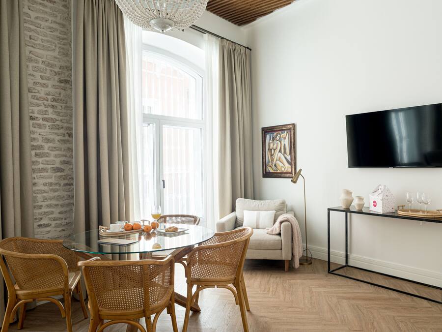 Een eetgedeelte met een glazen tafel, rieten stoelen, een witte bank en een televisie op een zwarte standaard.