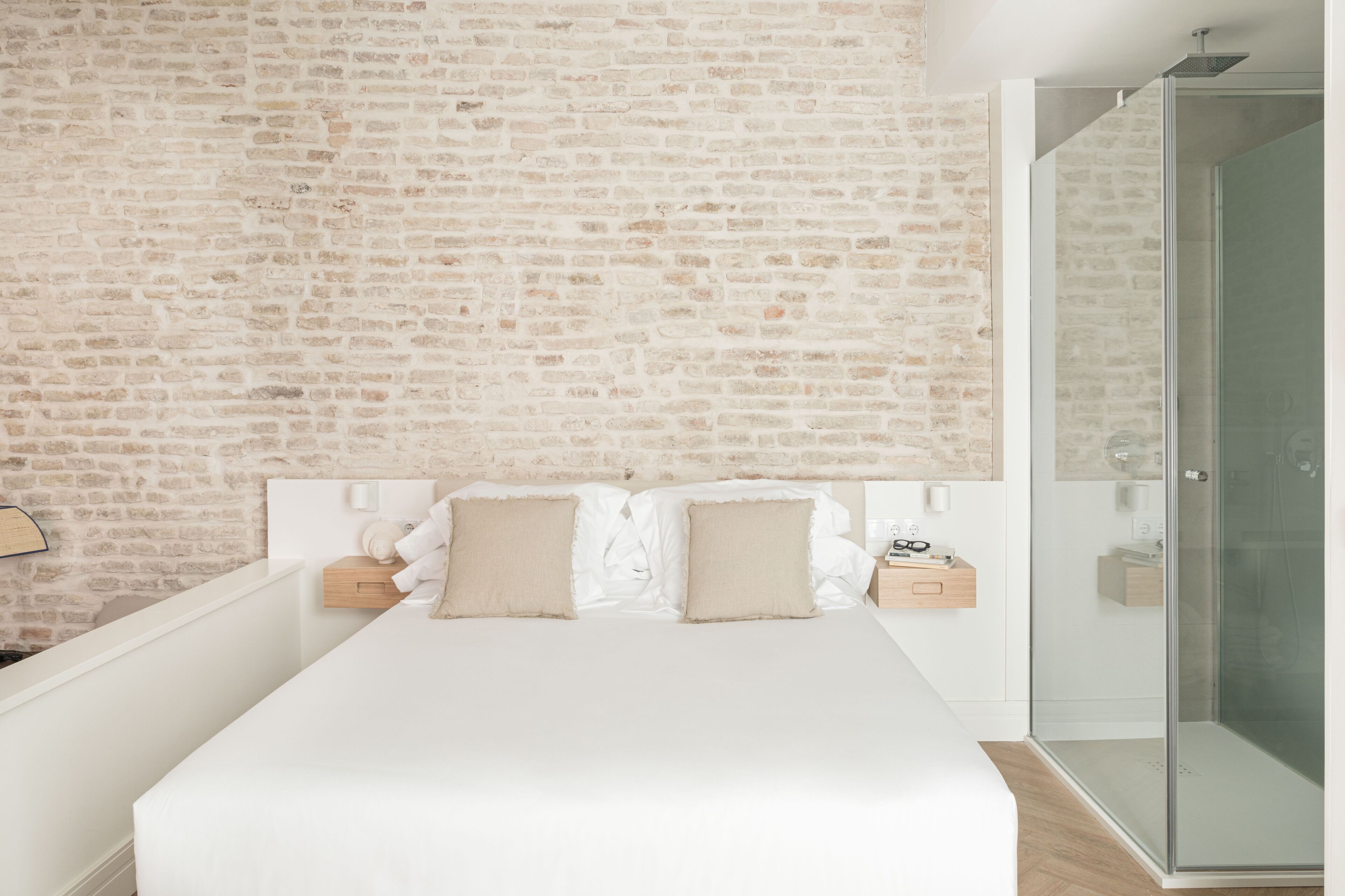 A modern bedroom with a brick-patterned wall, a large bed with white bedding, and a glass-enclosed shower area.