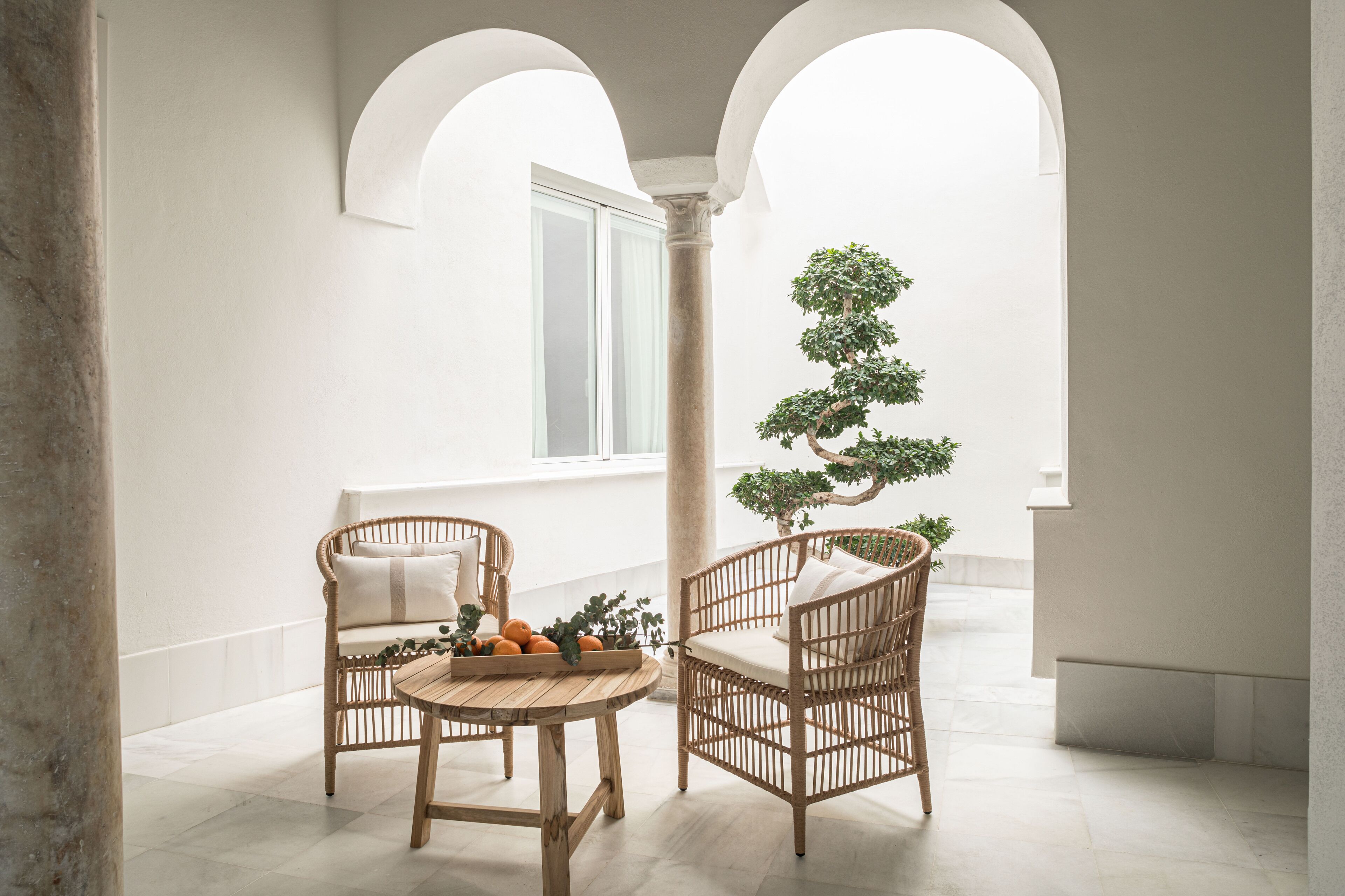 Un espace intérieur paisible, avec deux fauteuils en osier, une table en bois et un bonsaï, sous un plafond blanc orné de fenêtres en arc de cercle.