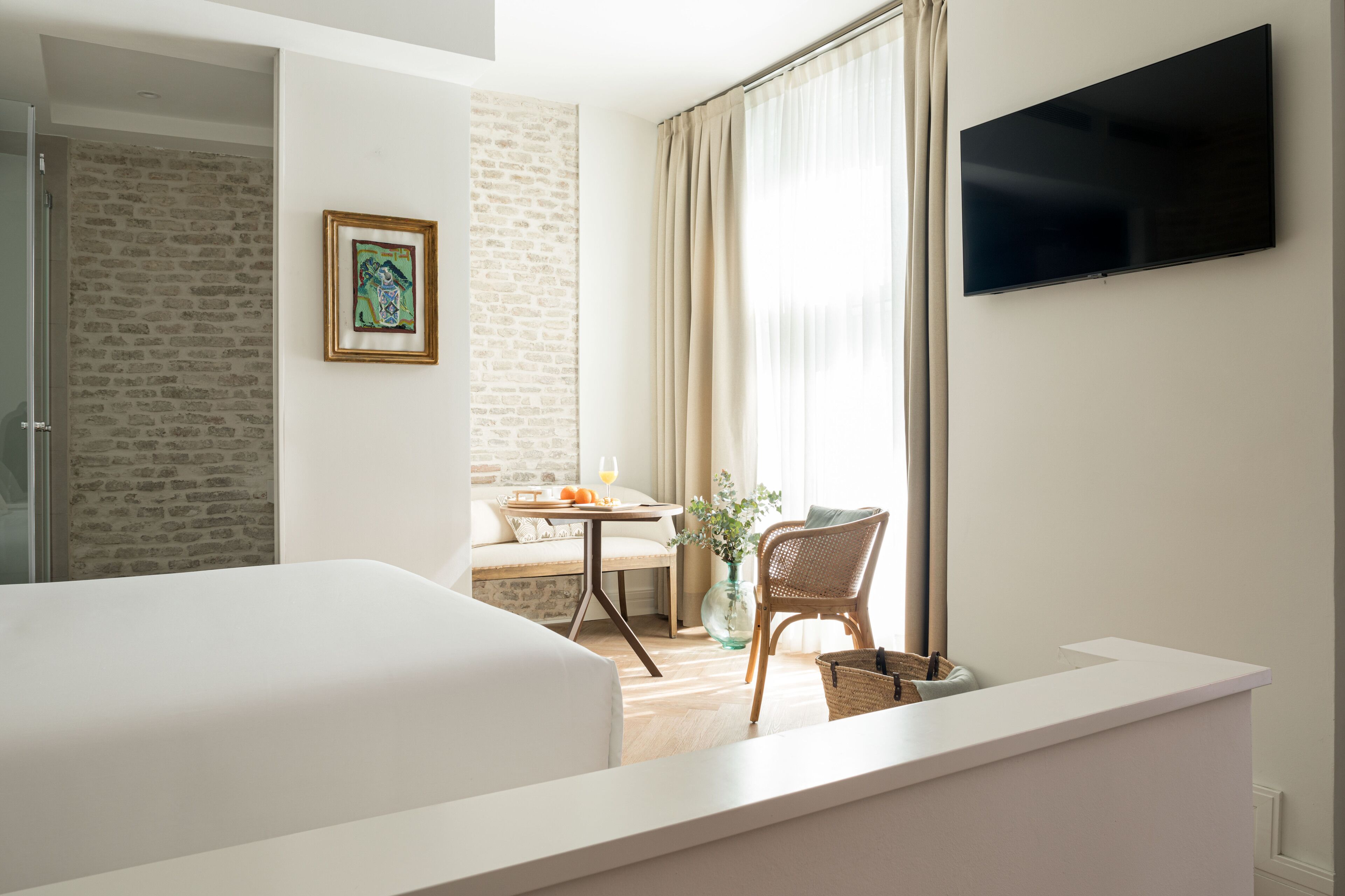 Une chambre d’hôtel avec un grand lit, un téléviseur fixé au mur, un coin repas avec une table et des chaises, et un mur en briques.