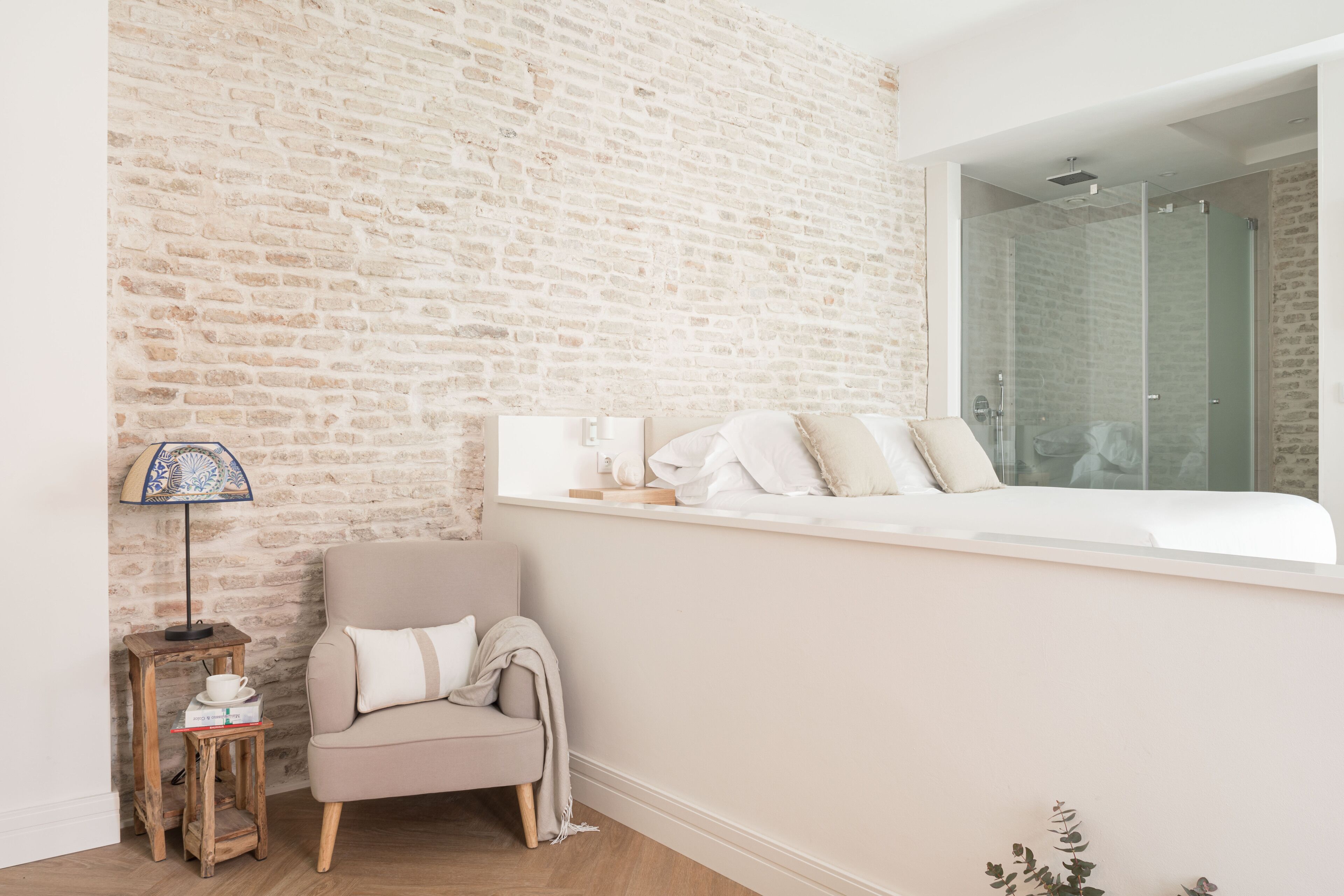 A bedroom with a brick wall, a window with a view of a shower, a chair, a small table with a lamp, and a bedside table with a cup and a book.