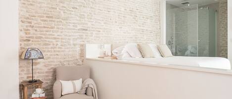 A bedroom with a brick wall, a window with a view of a shower, a chair, a small table with a lamp, and a bedside table with a cup and a book.