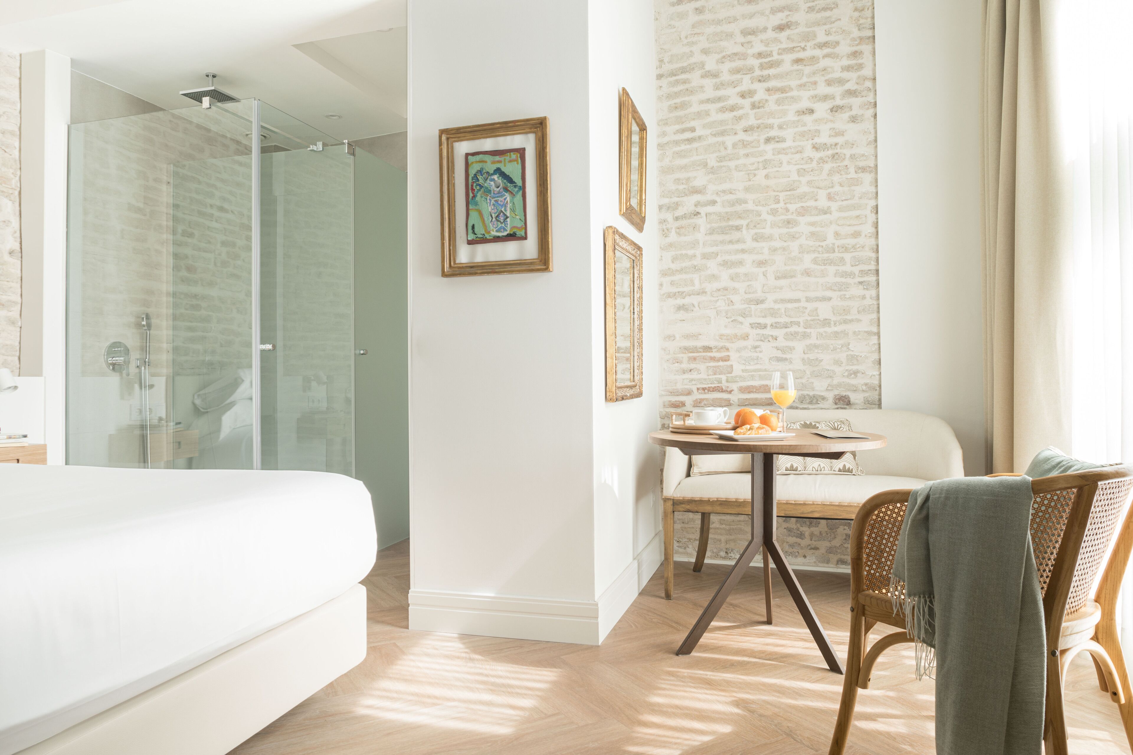 A modern hotel room with a glass shower, a bed, a dining area with a table and chairs, and artwork on the walls.