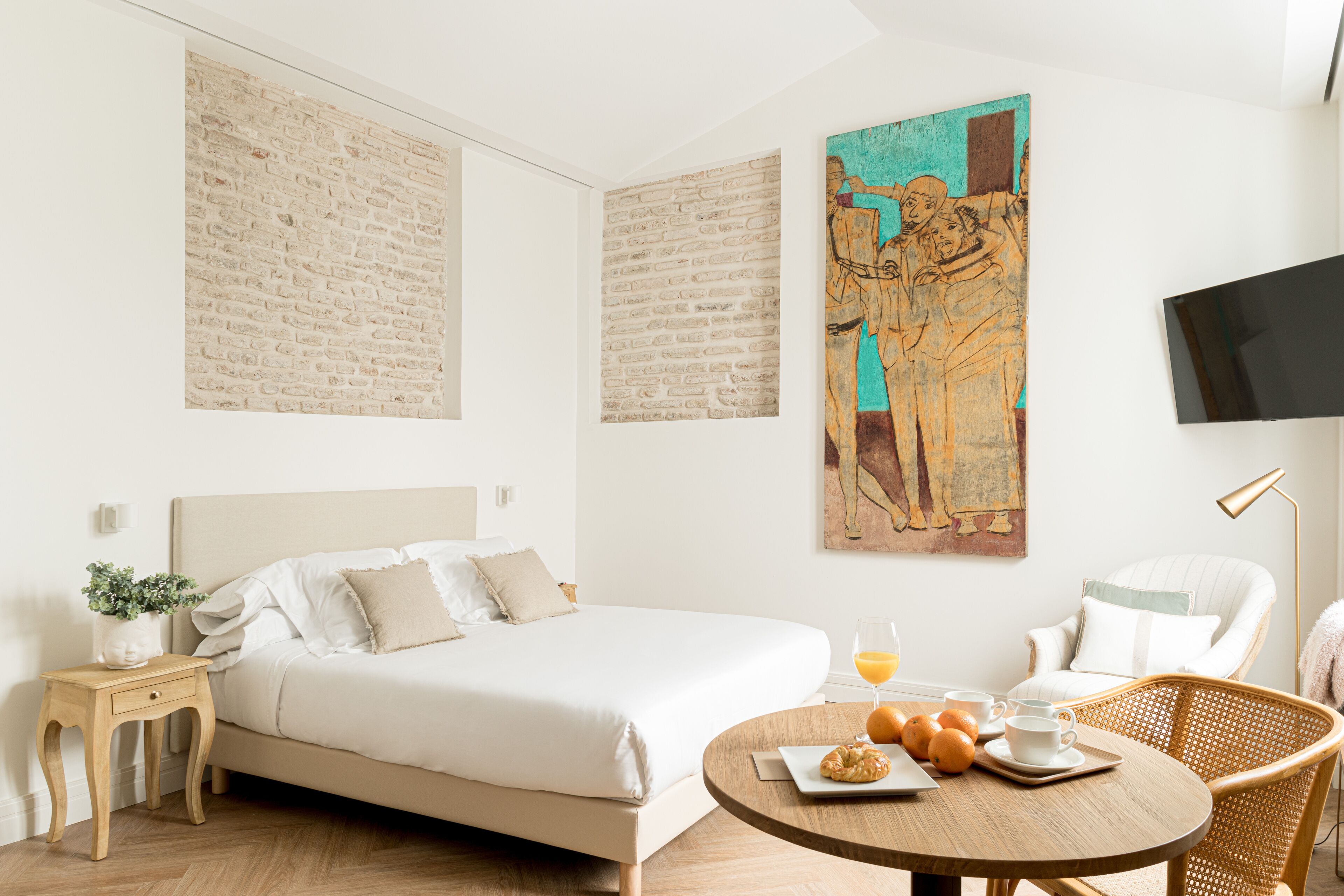 A modern hotel room with a large bed, a round dining table with breakfast items, a wicker chair, a flat-screen TV, and artwork on the walls.