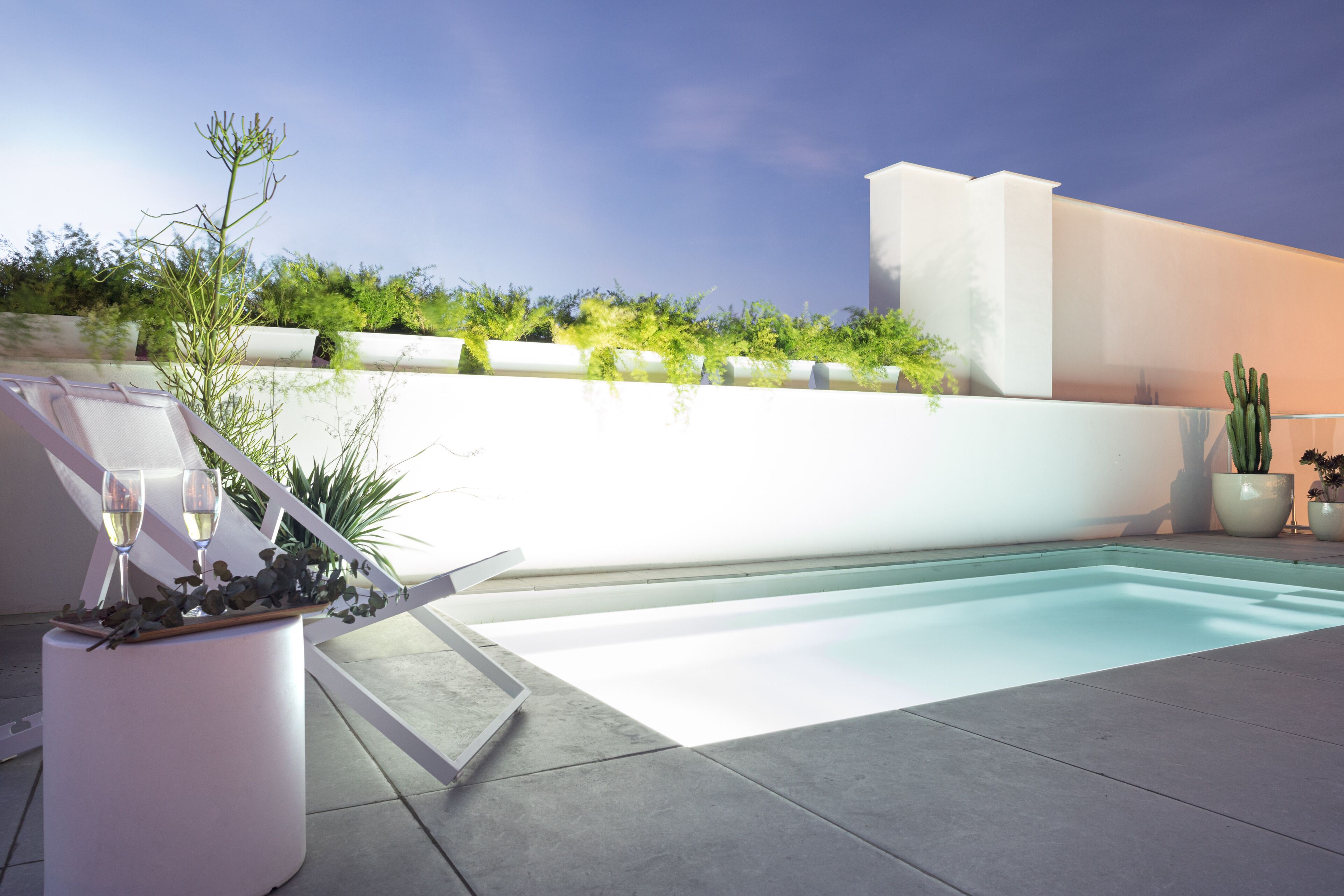 A modern outdoor pool area with a white wall and greenery, a lounge chair, and two wine glasses on a table.