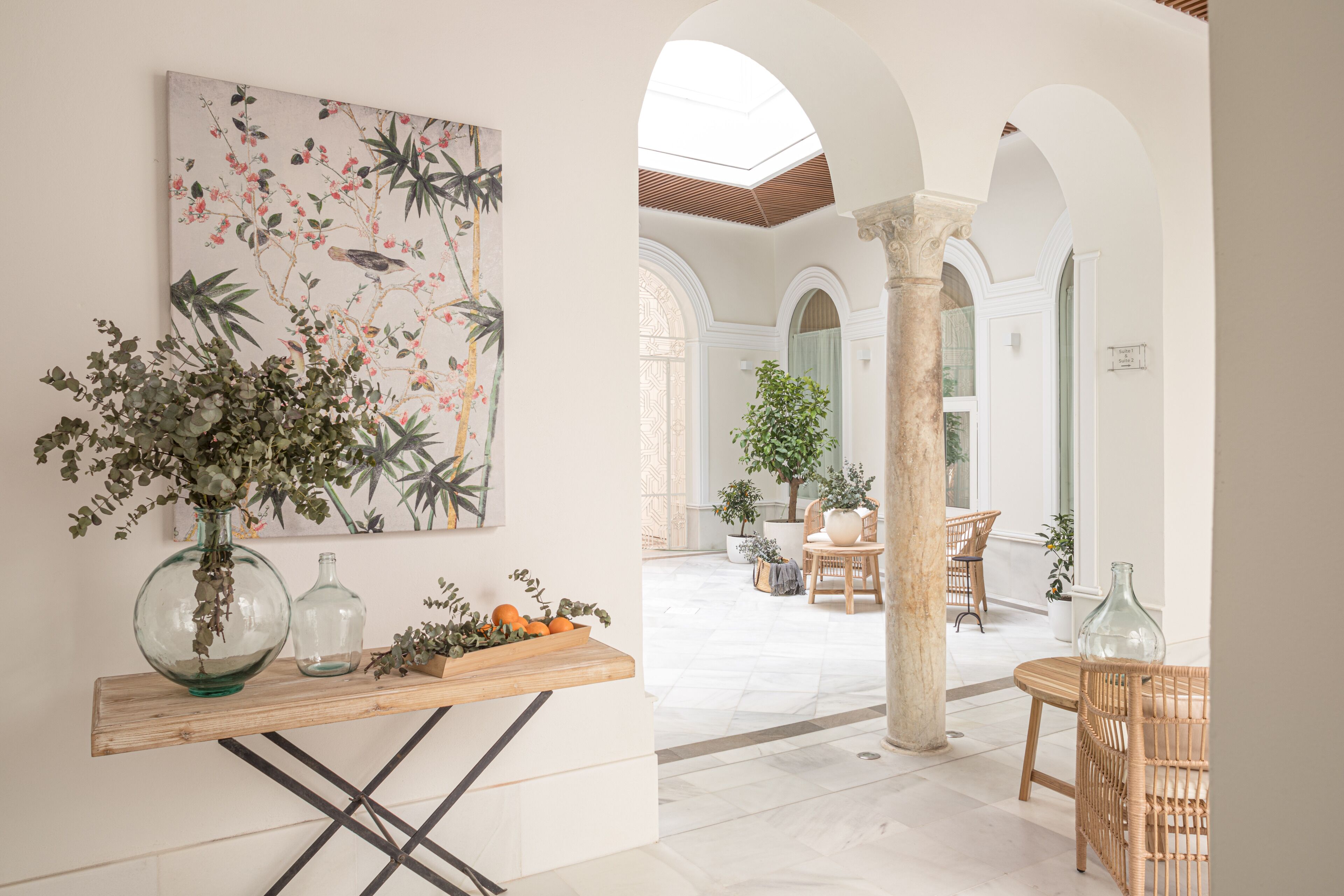 Amplo interior com mesa de madeira, uma pintura grande e vista para um pátio exterior com cadeiras e plantas.