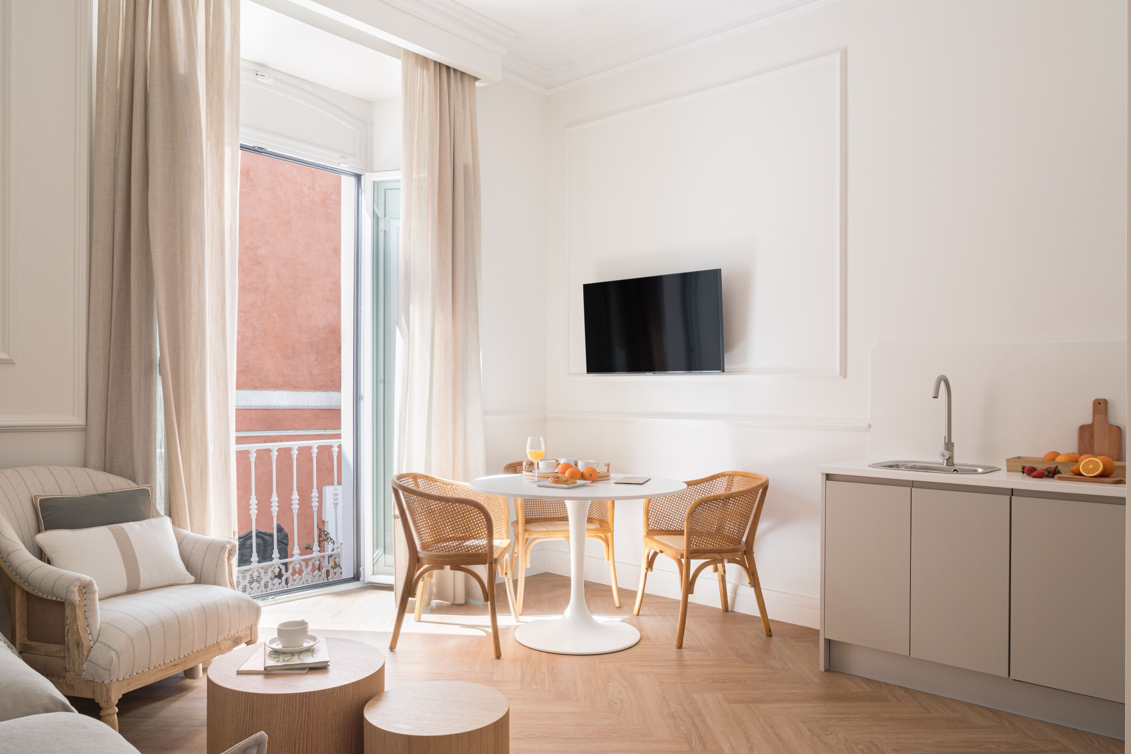 Una sala moderna con un sofá, una mesa de comedor con sillas, un televisor montado en la pared y una zona de cocina con un fregadero y fruta en la encimera.