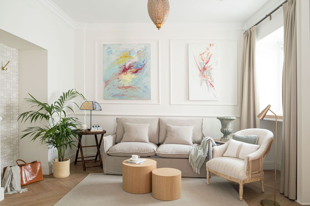 A living room with a beige sofa, two round wooden side tables, a floor lamp, a potted plant, and abstract wall art.