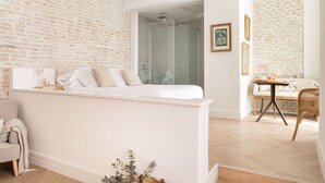 A modern hotel room with a white bed, a glass shower, a dining area with a table and chairs, and a brick wall. - MYLU SUITES by PUERTA CATEDRAL (Seville)