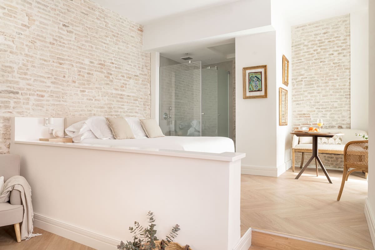 A modern hotel room with a white bed, a glass shower, a dining area with a table and chairs, and a brick wall.