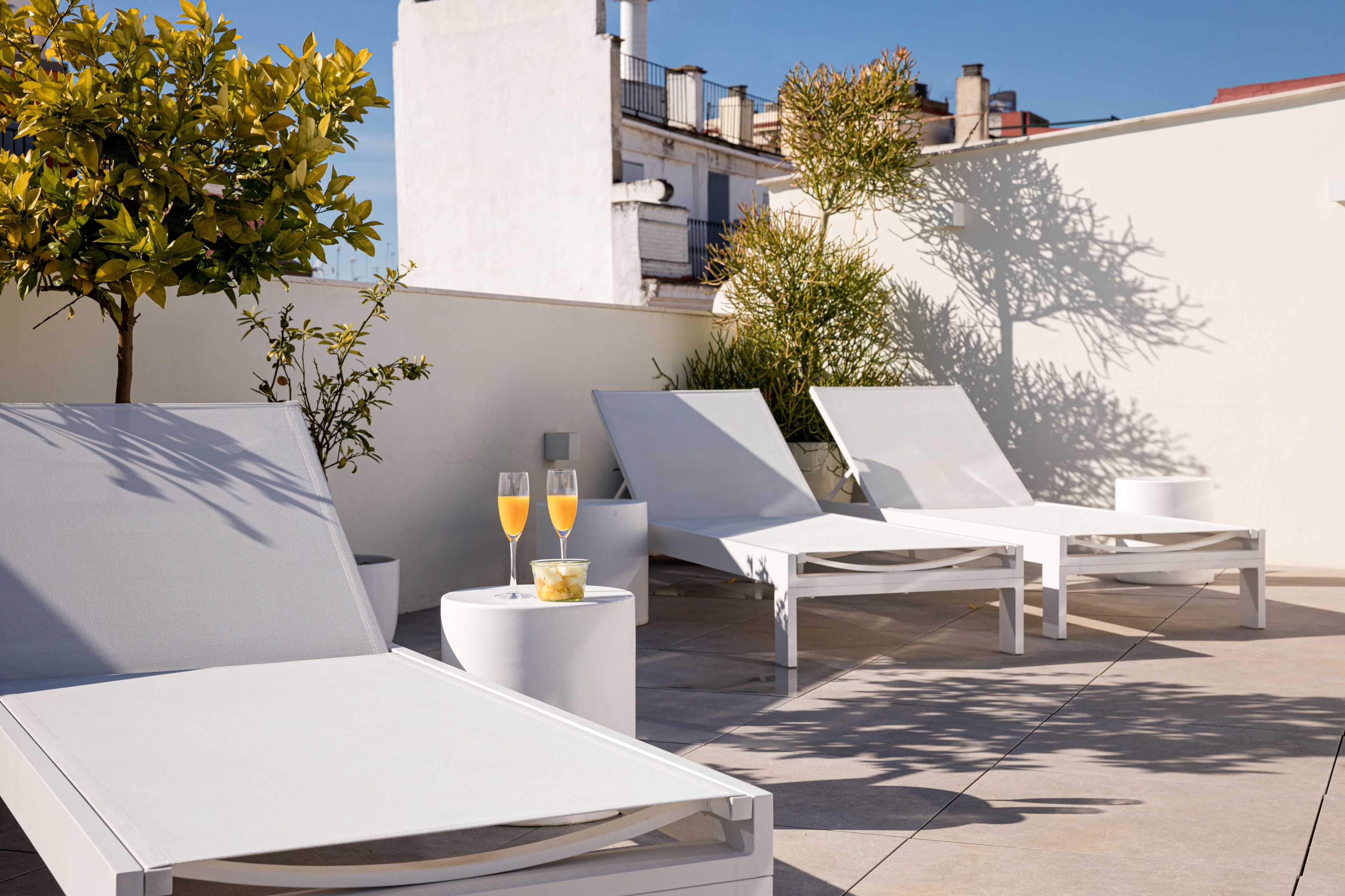 A rooftop terrace with white lounge chairs, a small table with two glasses of orange juice, and potted plants.