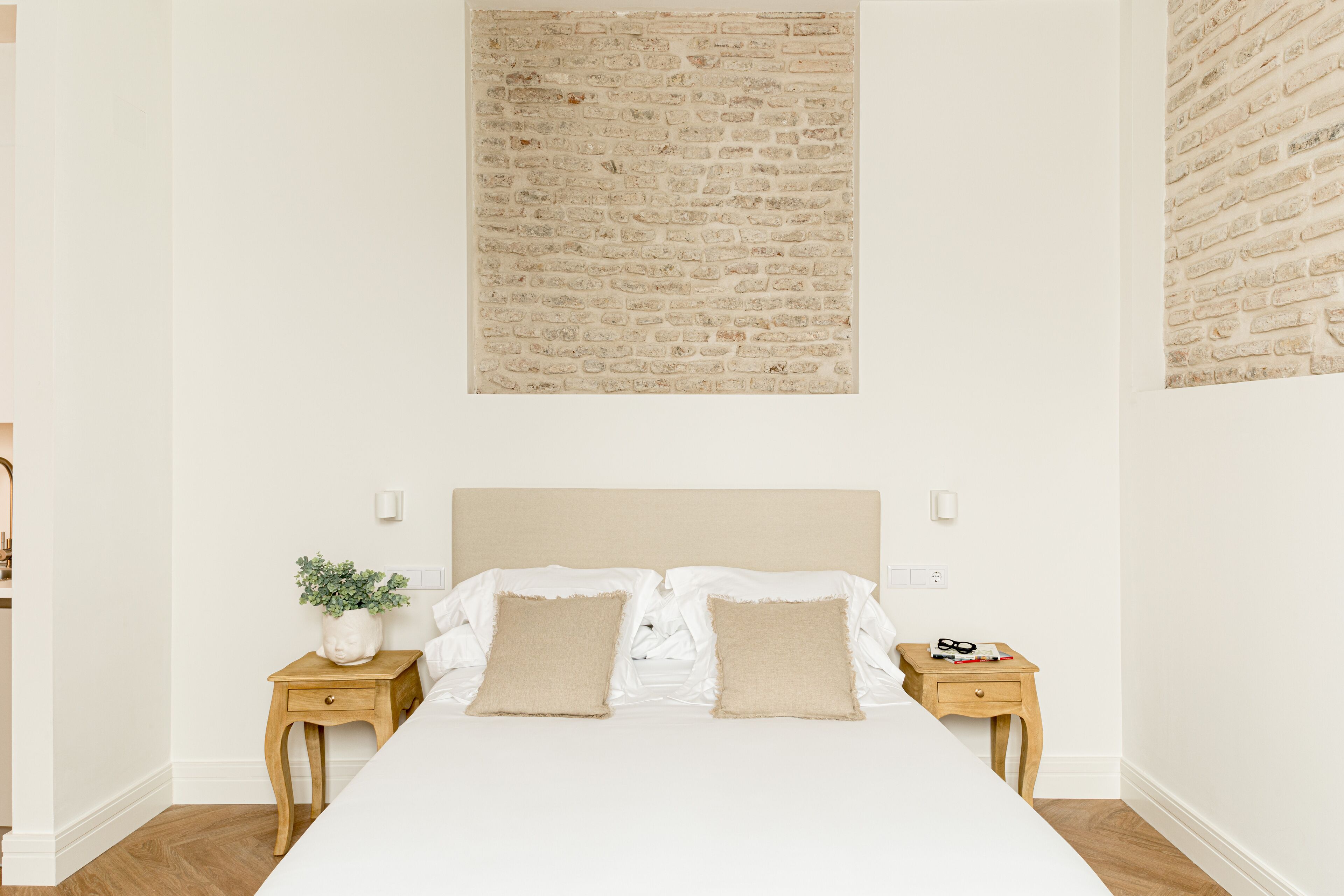 Un dormitorio con una cama grande, dos mesitas de noche, un muro con diseño de ladrillo y una pequeña planta en maceta.