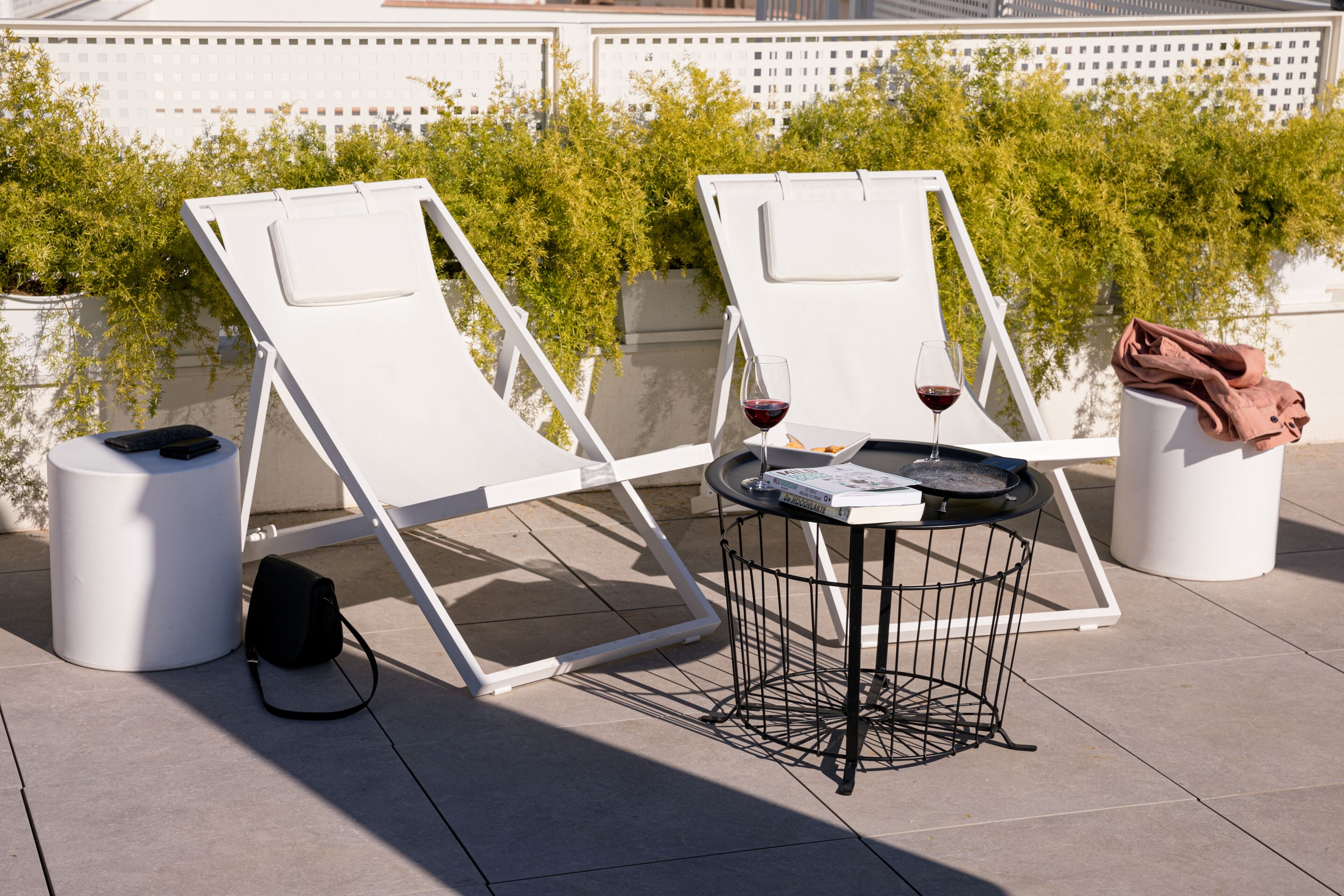 Två vita loungestolar med ett bord och ett glas rött vin mellan sig, placerade på en takterrass med krukväxter och ett vitt staket i bakgrunden.