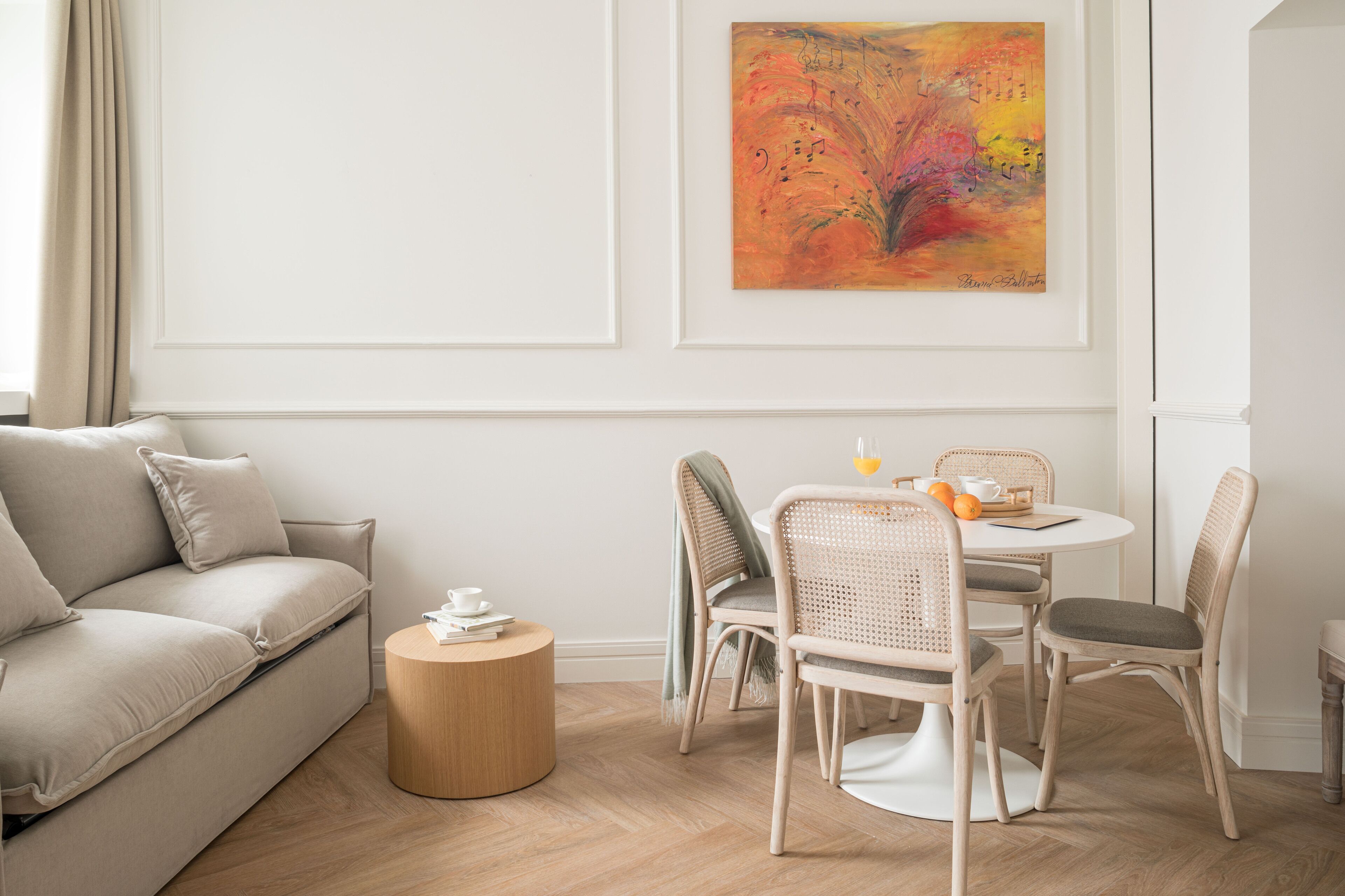 Un salon moderne avec un canapé beige, une table basse ronde en bois, des chaises en osier et une table à manger agrémentée d’un saladier de fruits et d’une tasse. Un tableau abstrait aux couleurs vives orne le mur.