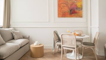 A modern living room with a beige sofa, a round wooden coffee table, wicker chairs, and a dining table with a fruit bowl and a cup. A colorful abstract painting is on the wall.