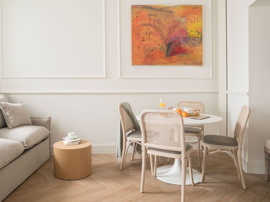 A modern living room with a beige sofa, a round wooden coffee table, wicker chairs, and a dining table with a fruit bowl and a cup. A colorful abstract painting is on the wall.