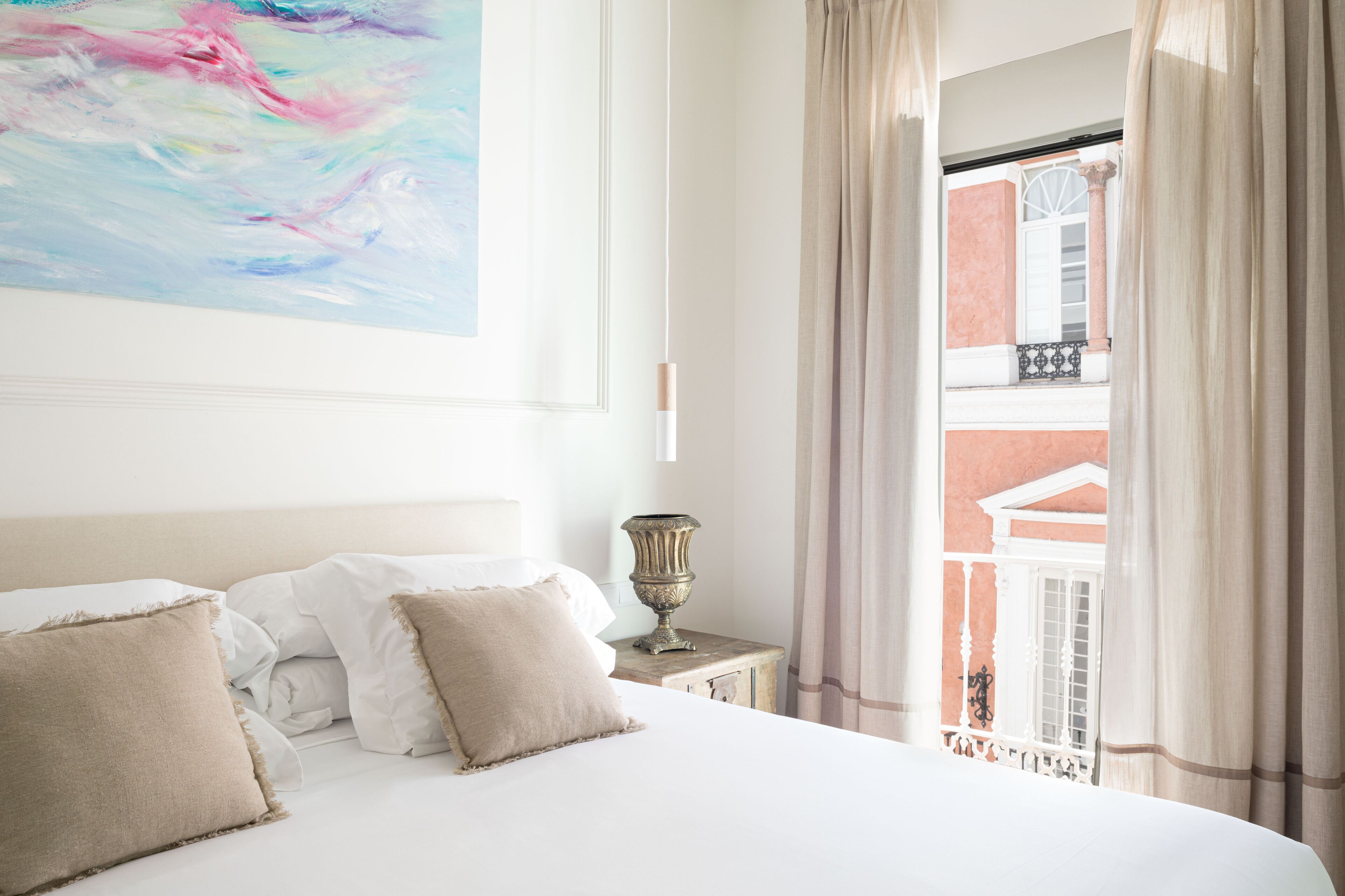 A hotel room with a bed, pillows, a bedside table with a lamp, and a view of the outside balcony.