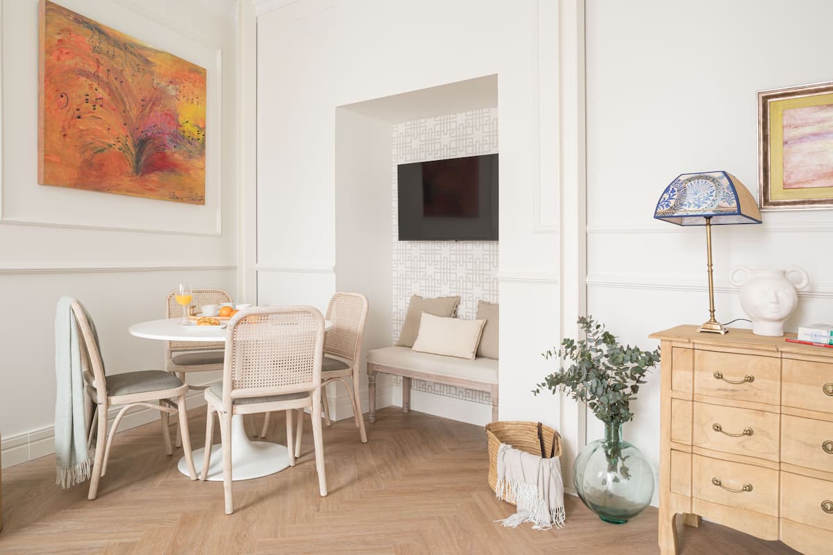 A dining area with a round table, wicker chairs, a bench, a vase with greenery, a lamp, and a television on the wall.