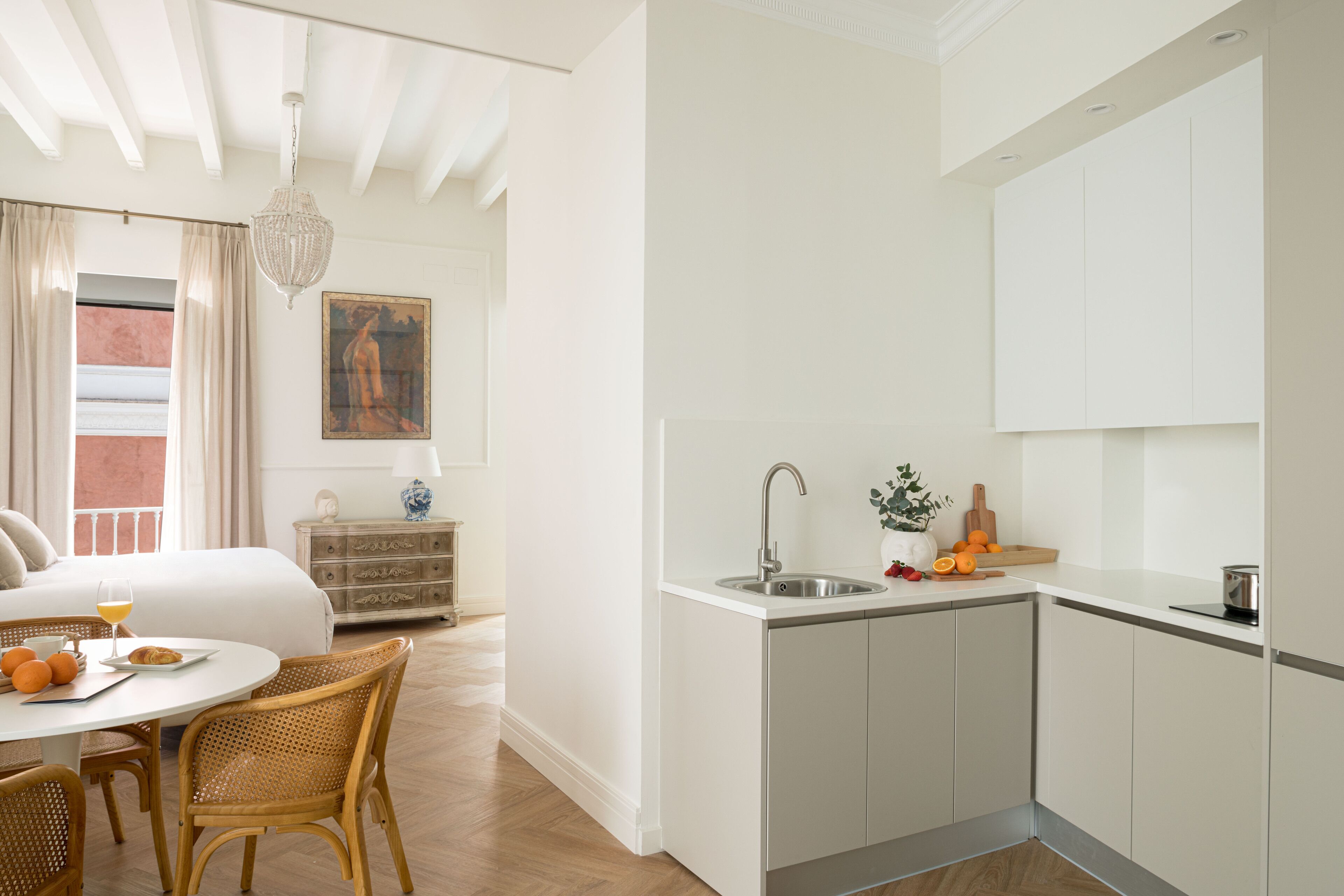 A modern hotel room with a bed, breakfast table, wicker chairs, a dining area, a kitchen with a sink, and a painting on the wall.