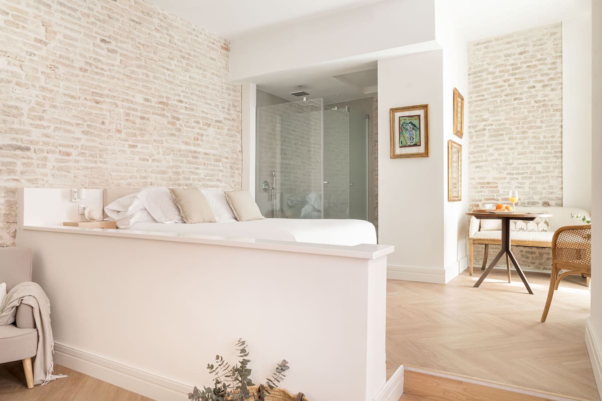 A modern hotel room with a brick wall, a glass shower, a white bed, a small table with a chair, and a wicker chair.