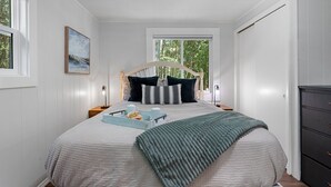 Une chambre bien rangée, avec un grand lit, une table de chevet avec une lampe, une commode et une fenêtre donnant sur des arbres.