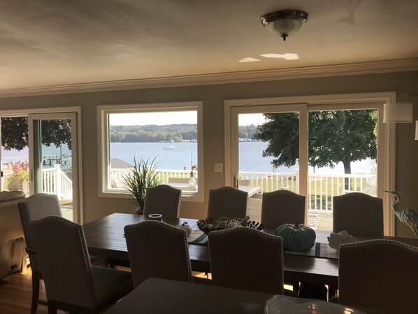 A dining area with a view of a lake and dock, featuring a wooden table, chairs, and a potted plant.