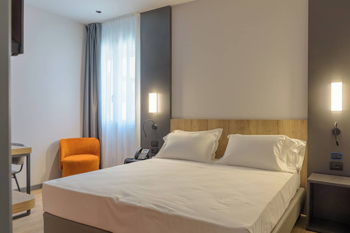 Una habitación de hotel con una cama, una silla naranja, un escritorio, un teléfono y una mesita de noche.