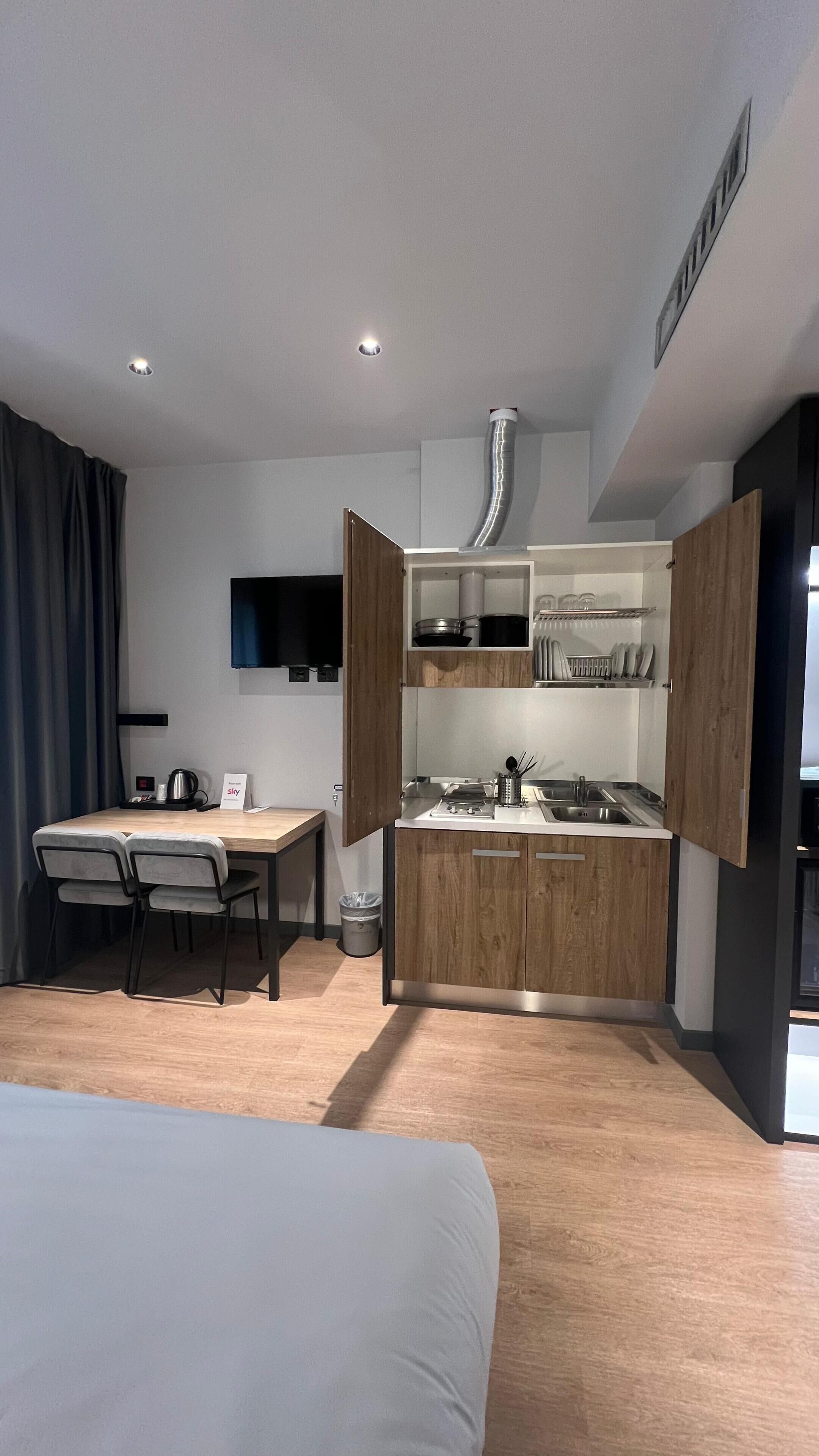 A compact hotel room with a bed, a dining table with chairs, a kitchenette, and a TV mounted on the wall.