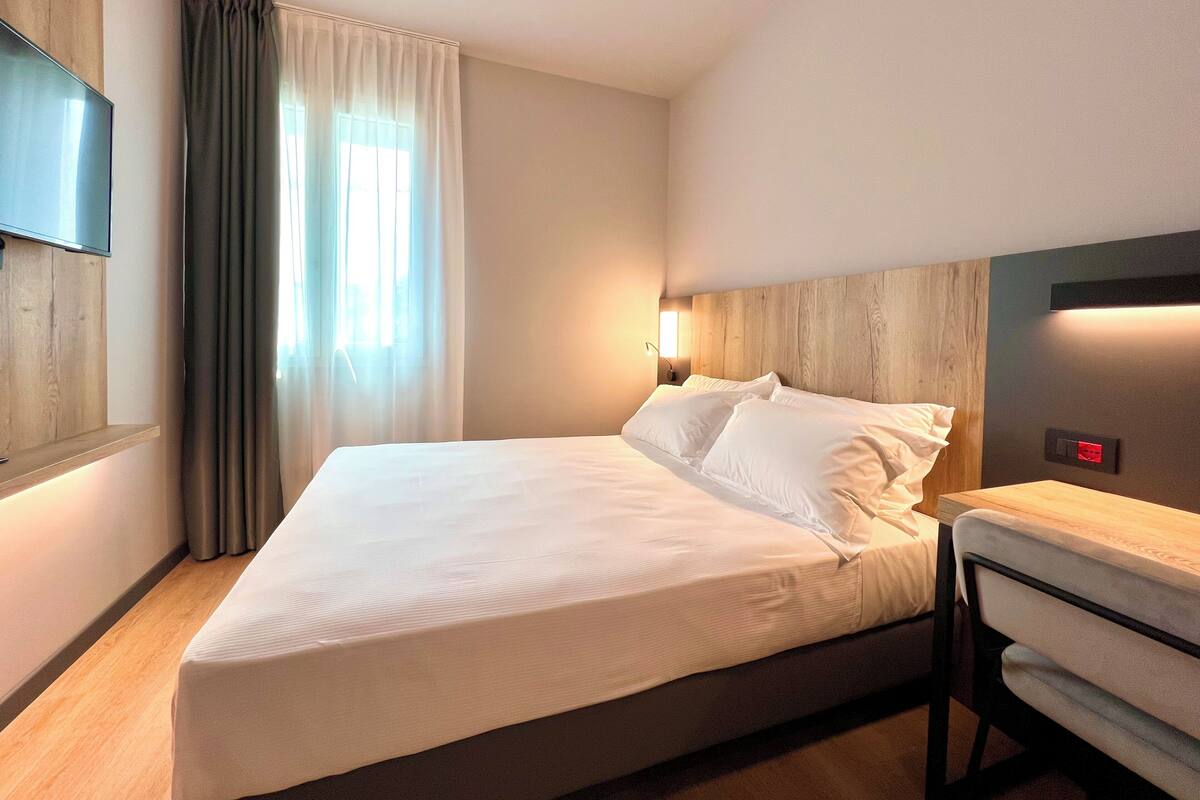Una habitación de hotel con cama, televisor montado en la pared, mesita de noche y ventana con cortinas.